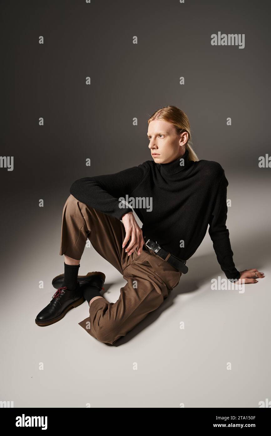 Gut aussehende, nicht binäre Person in schwarzem Rollkragen und brauner Hose, die auf dem Boden sitzt, Mode Stockfoto