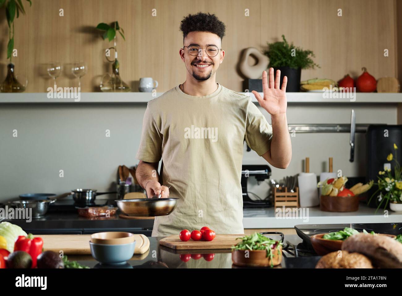 Mittelgroße Aufnahme des lächelnden jungen Küchenchefs aus dem Nahen Osten, der glücklich in der Kamera winkt, während er in der Küche posiert, im Kopierraum Stockfoto