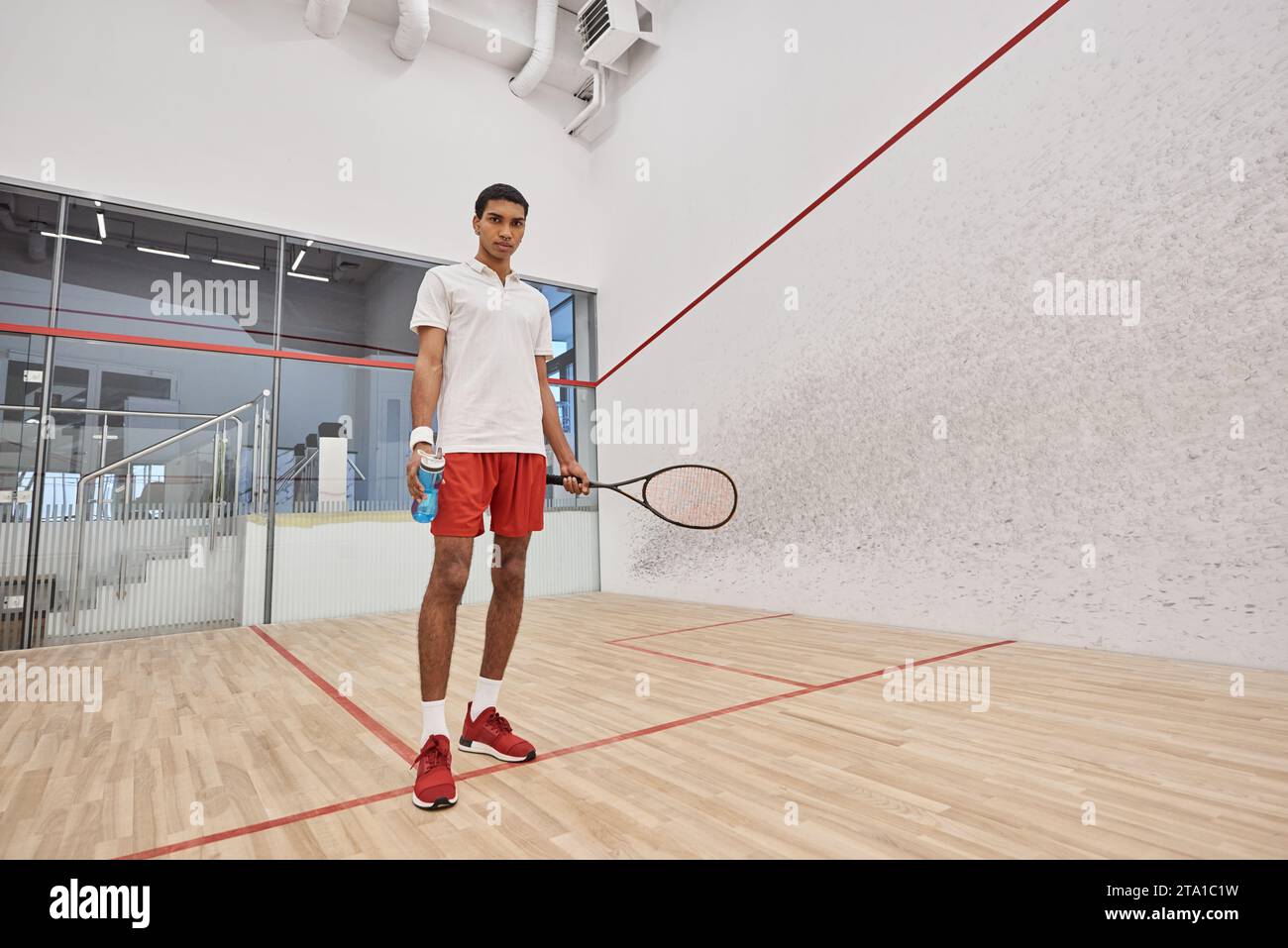 Junger afroamerikanischer Mann, der nach dem Spielen eine Sportflasche mit Wasser und Squash-Schläger hält Stockfoto