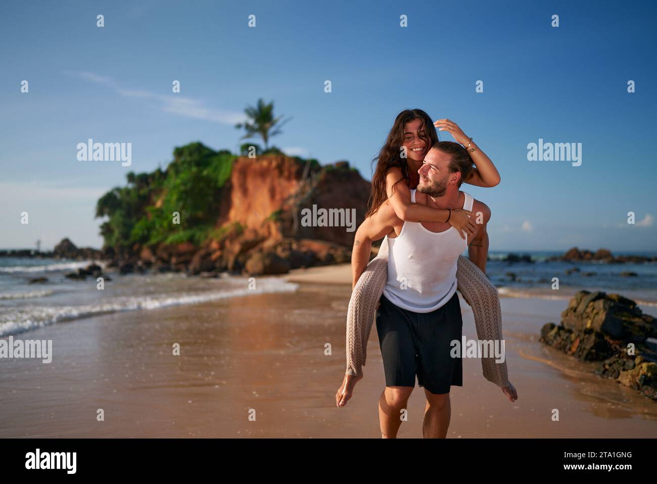 Glückliches Paar genießt die Flitterwochen am tropischen Strand. Der Mann trägt eine Frau mit Huckepack vor Freude. Barfuß lächelt, spaziert entlang des Ufers. Liebe, romantische Blüte Stockfoto