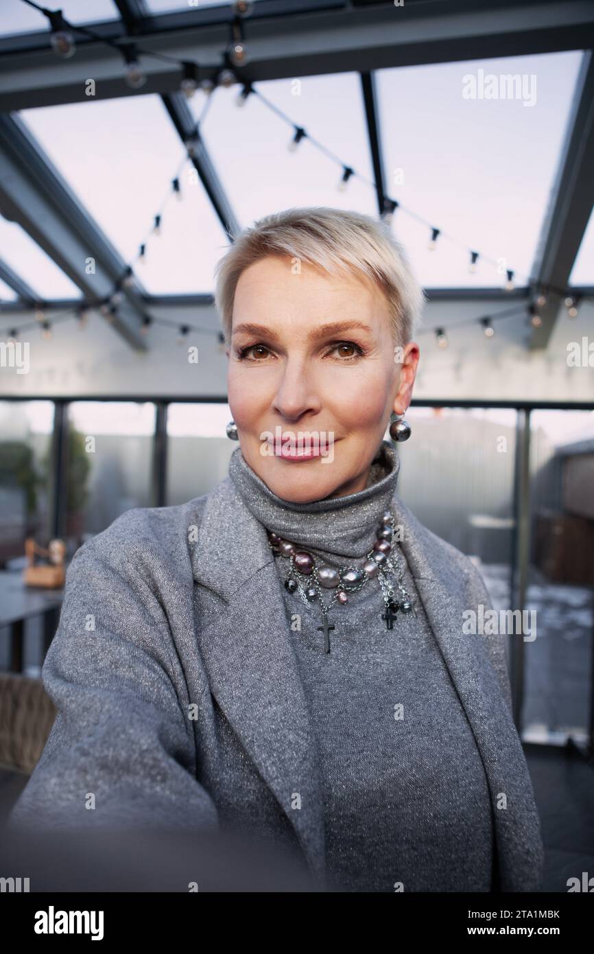 Selfie-Aufnahme einer schönen Reifen blonden Frau mit kurzen Haaren, die elegante graue Kleidung mit Accessoires trägt. Nahaufnahme Porträt von elegantem Mittelteil Stockfoto