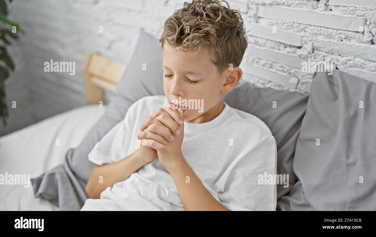 Der entzückende blonde Junge sitzt in einem gemütlichen Bett und entspannt sich am frühen Morgen mit Glauben. In eine Decke gehüllt, färben Gebet und Hoffnung den Raum, während er tröstet Stockfoto