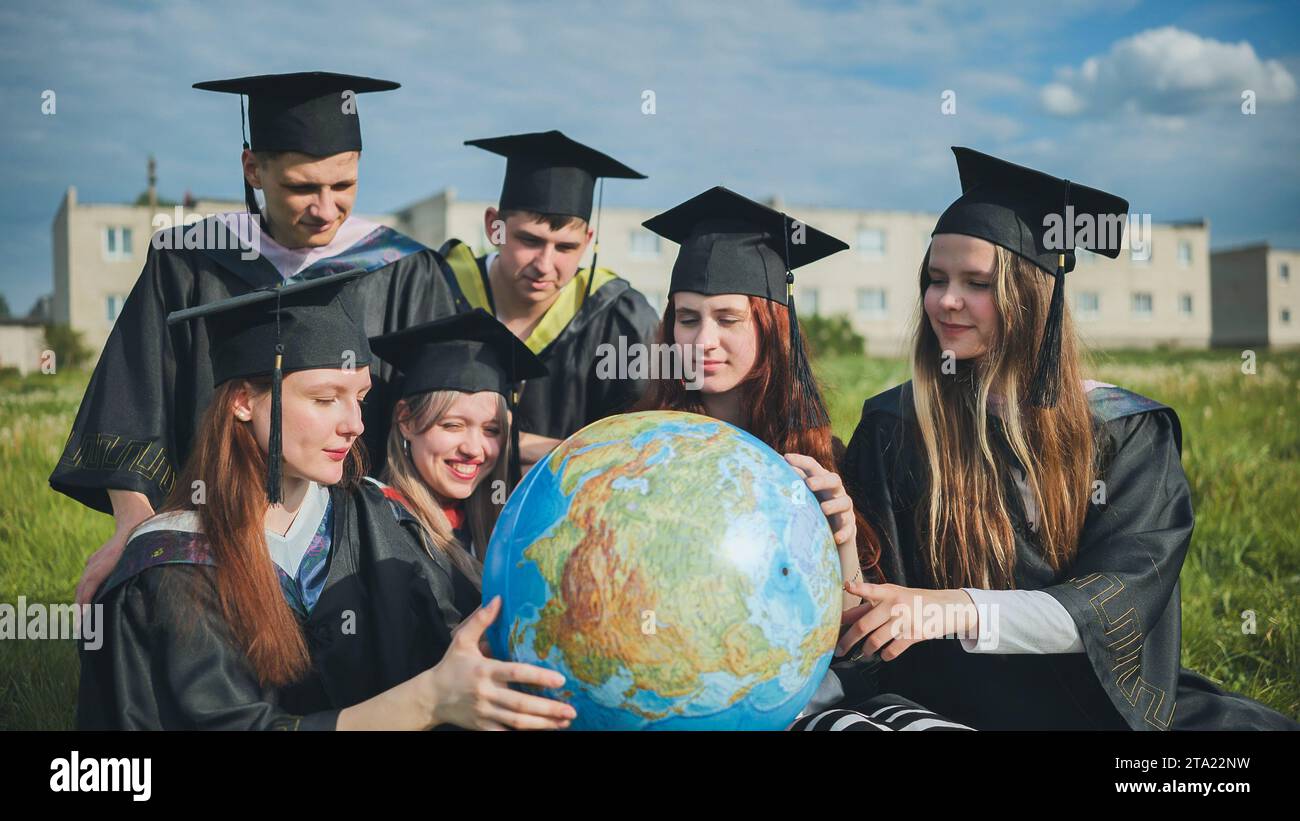 Absolventen in schwarzen Gewändern untersuchen einen geografischen Globus, der auf dem Gras sitzt. Stockfoto