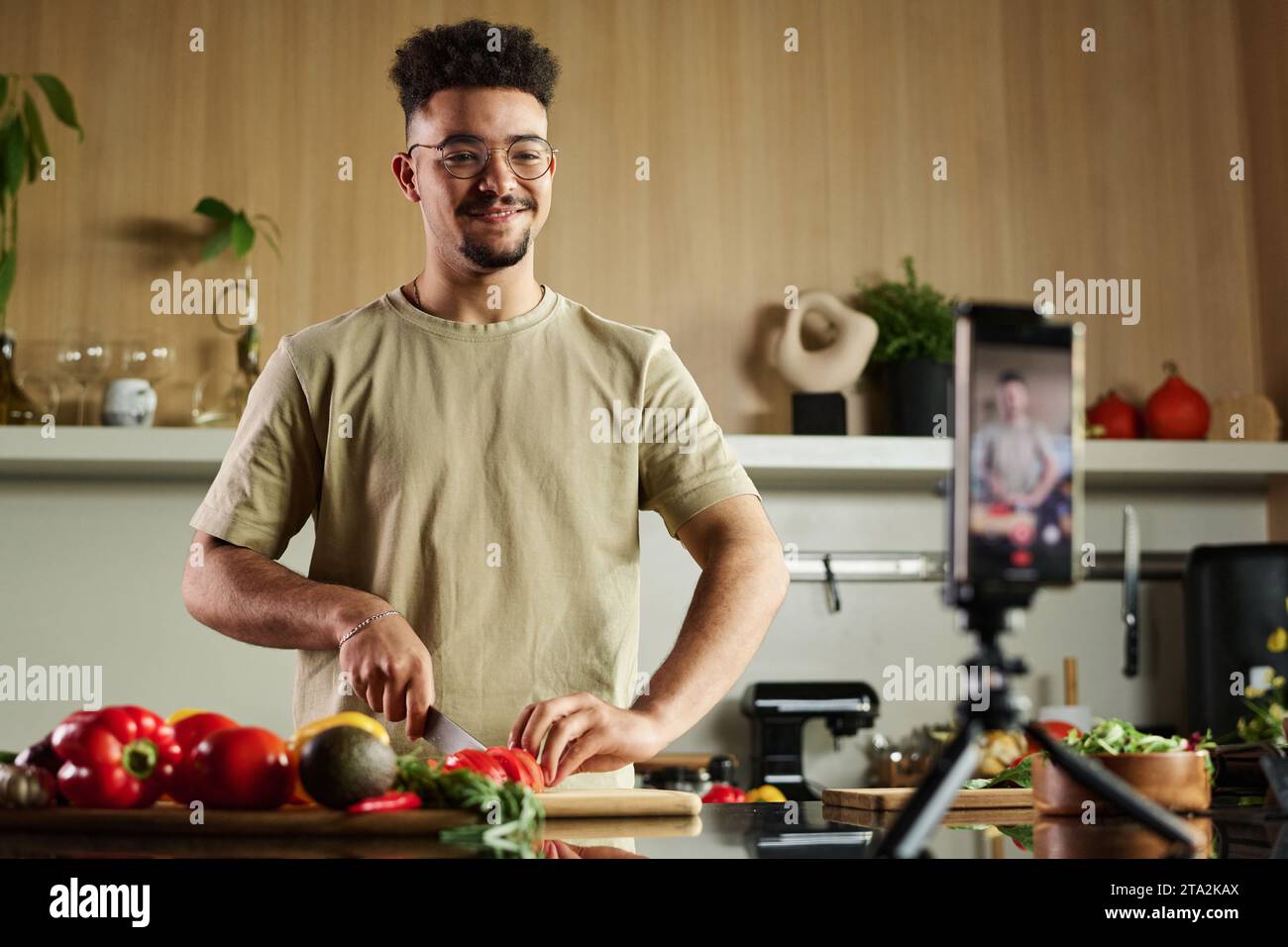 Mittlere Aufnahme eines jungen Koch aus dem Nahen Osten, der an der Küchentheke steht und Tomaten auf Schneidebrett schneidet, während er mit der Smartphone-Kamera gefilmt wird Stockfoto