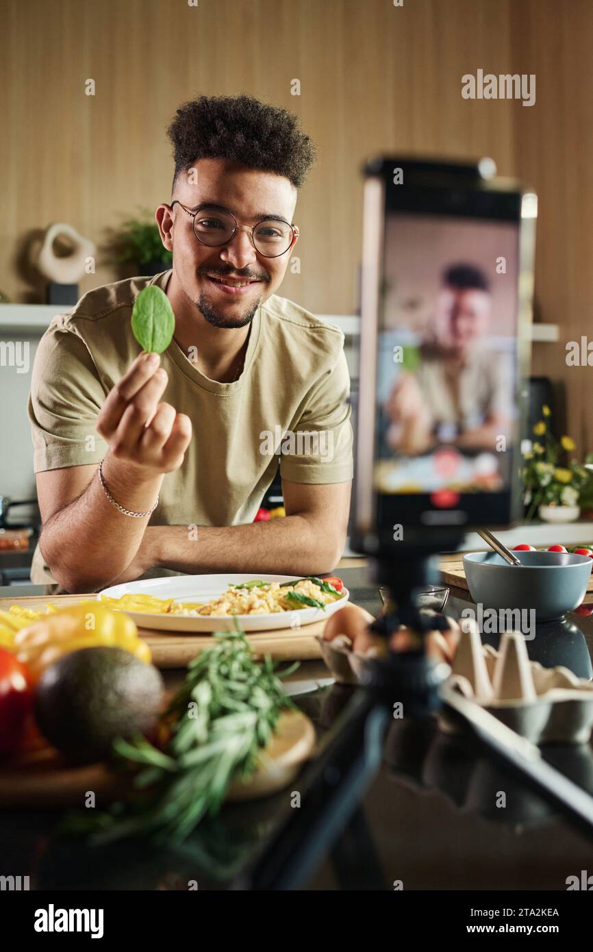 Porträt eines lächelnden jungen Bloggers aus dem Nahen Osten in der Küche, der Spinat auf der Smartphone-Kamera zeigt, um Videos für seinen Kanal zu machen Stockfoto