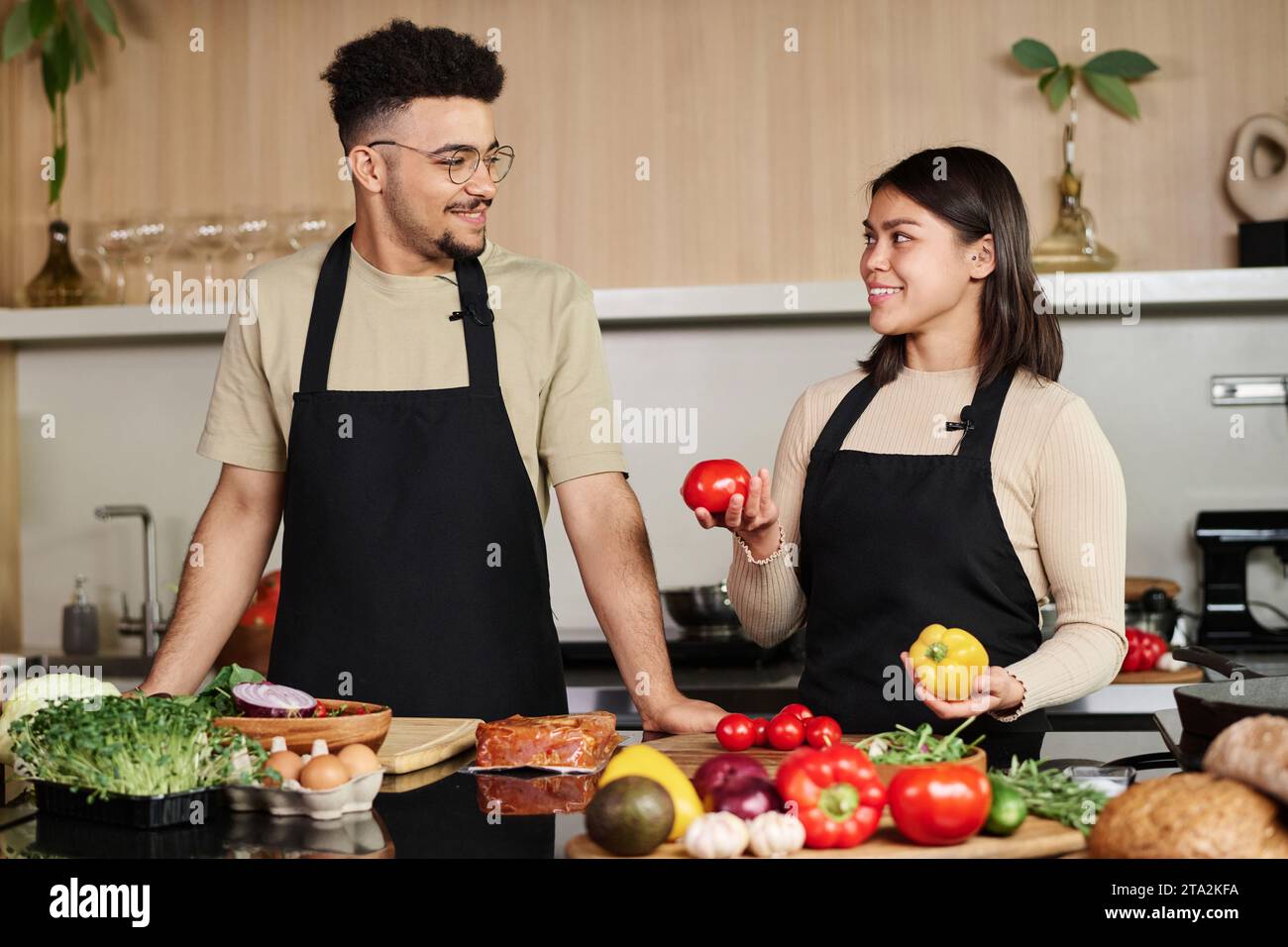 Junger Mann aus dem Nahen Osten und glückliches hispanisches Mädchen mit Gemüse in den Händen, die sich anschauen, während sie in der Küche posieren Stockfoto