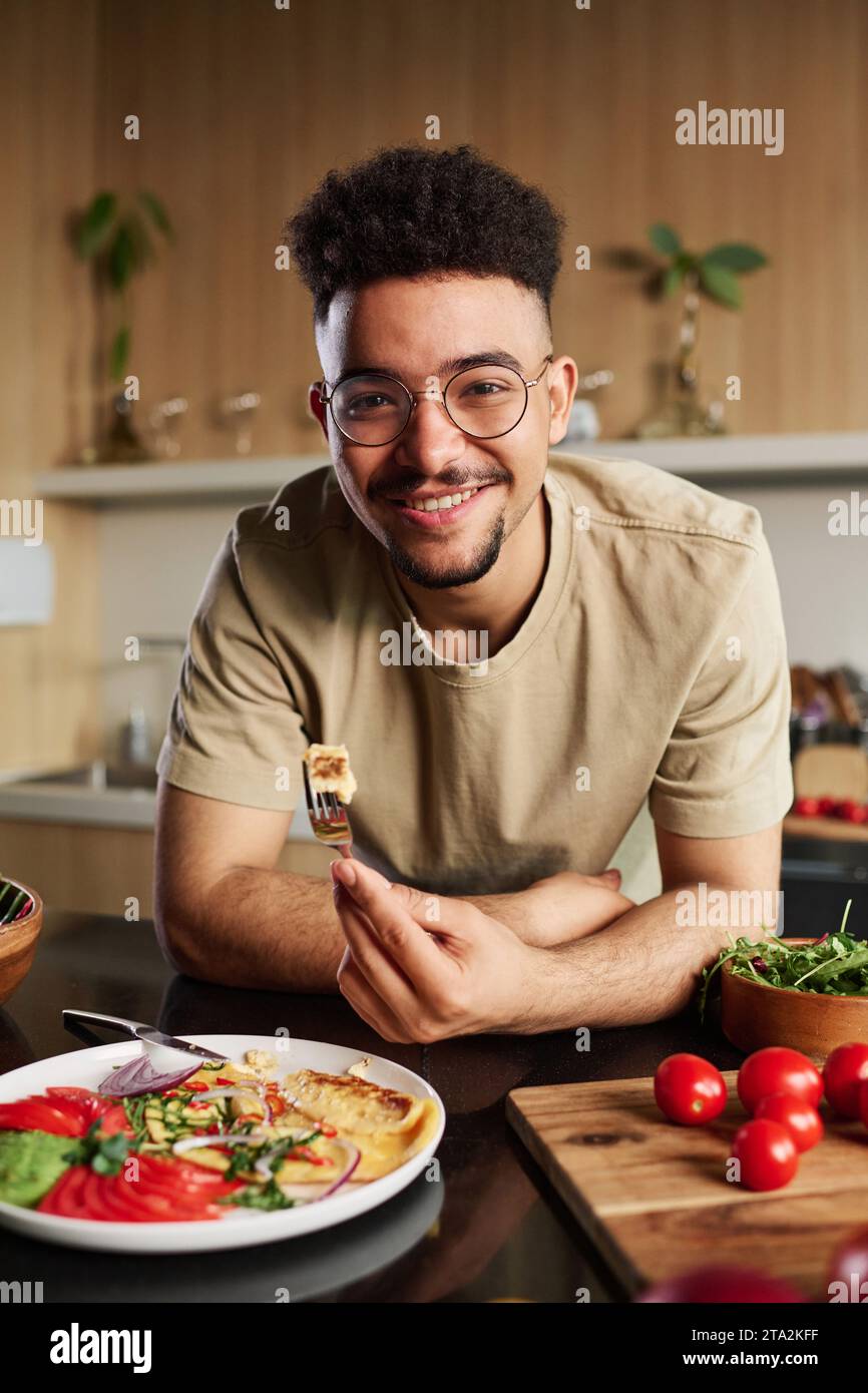 Junger Influencer aus dem Nahen Osten, der in die Kamera blickt und eine Gabel mit einem Stück Omelett auf dem Küchentisch mit Geschirr und Gemüse hält Stockfoto