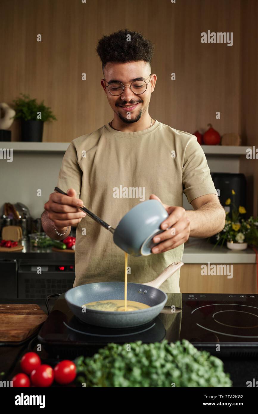 Mittlere vertikale Aufnahme des lächelnden jungen Küchenchefs aus dem Nahen Osten, der geschlagene Eier auf die Pfanne gießt, während er vor dem Küchenherd posiert Stockfoto