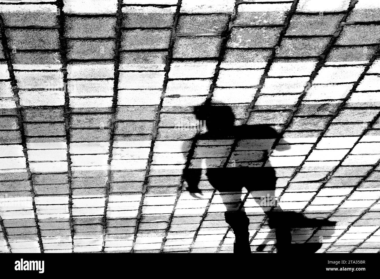 Schattensilhouette einer Person, die die Kopfsteinpflasterstraße auf Pelikanüberquerung überquert, in Schwarz-weiß Stockfoto