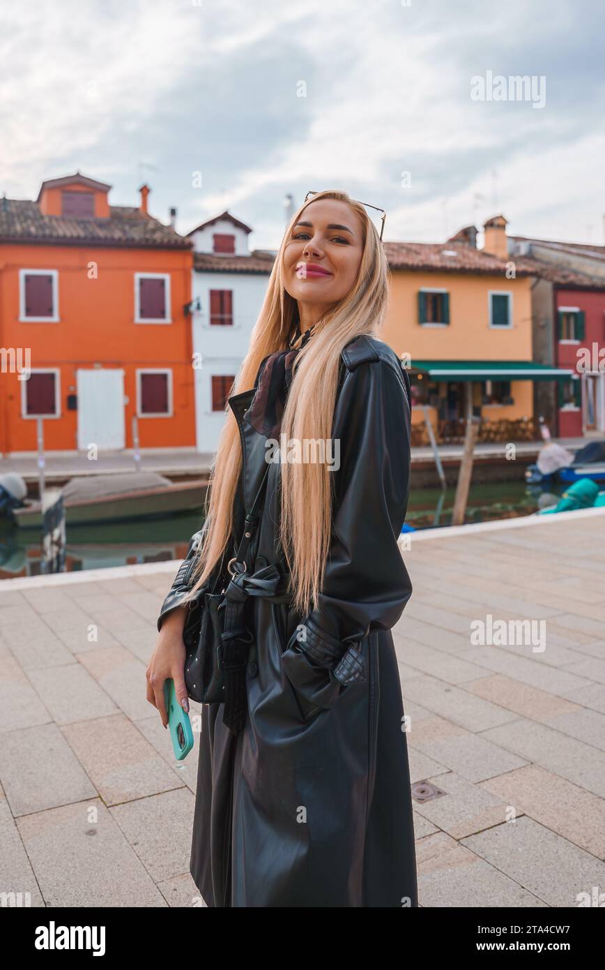Stilvolle Frau im schwarzen Ledermantel posiert vor bunten Gebäuden in Venedig, Italien Stockfoto