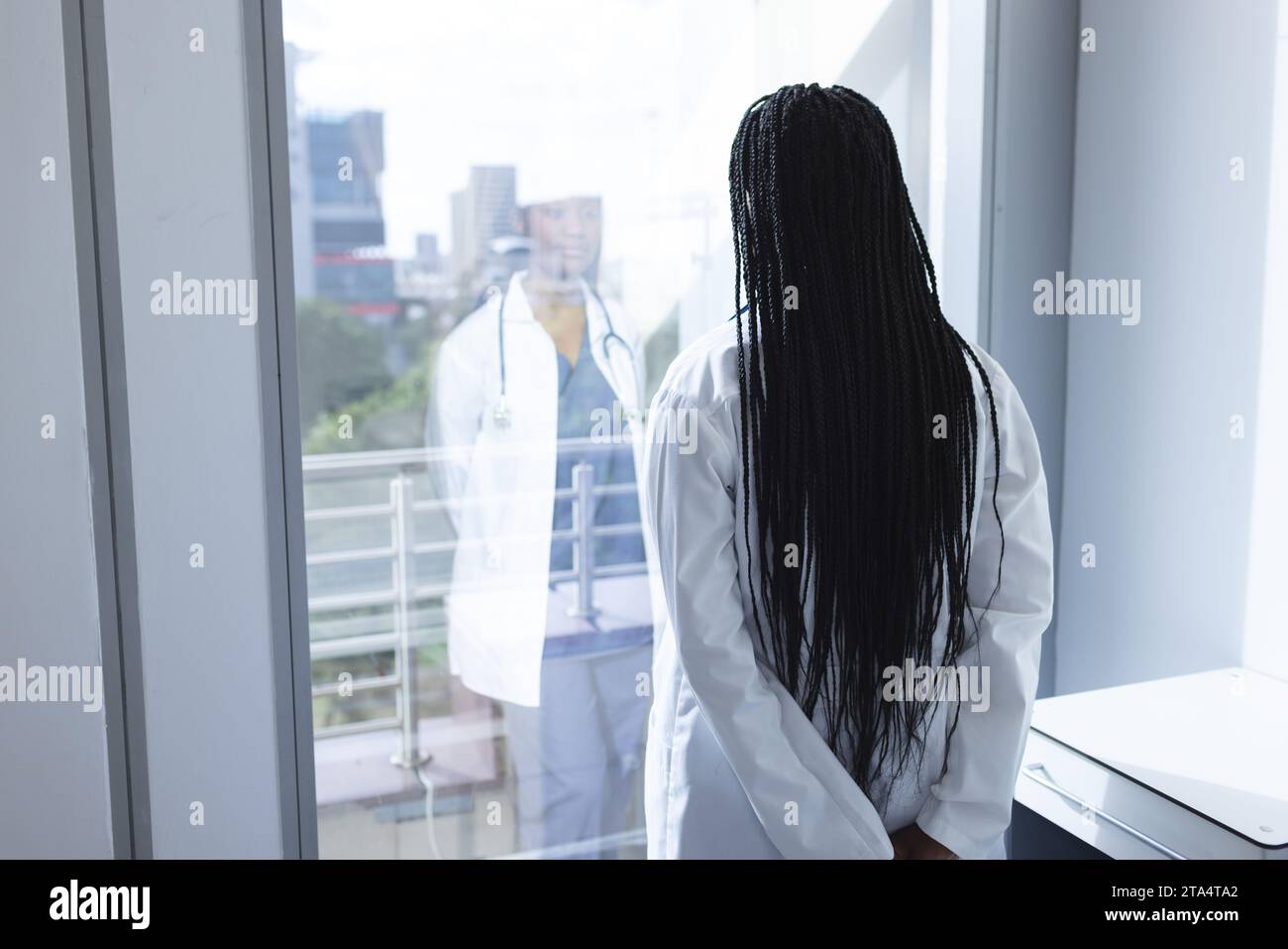 Nachdenkliche afroamerikanische Ärztin, die Laborkittel trägt und durch das Fenster im Krankenhauszimmer schaut Stockfoto