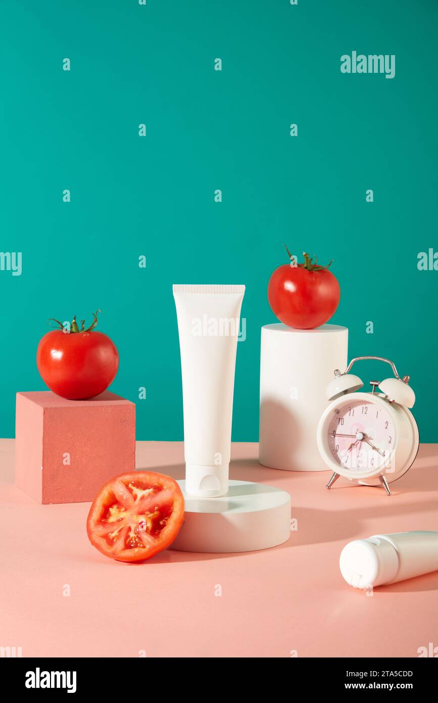 Ein unbeschriftetes Kosmetikröhrchen wird auf einem weißen Podium angezeigt. Zwei Tomaten werden auf Würfel neben einem Wecker gelegt. Türkisfarbener und pastellrosafarbener Backgrou Stockfoto