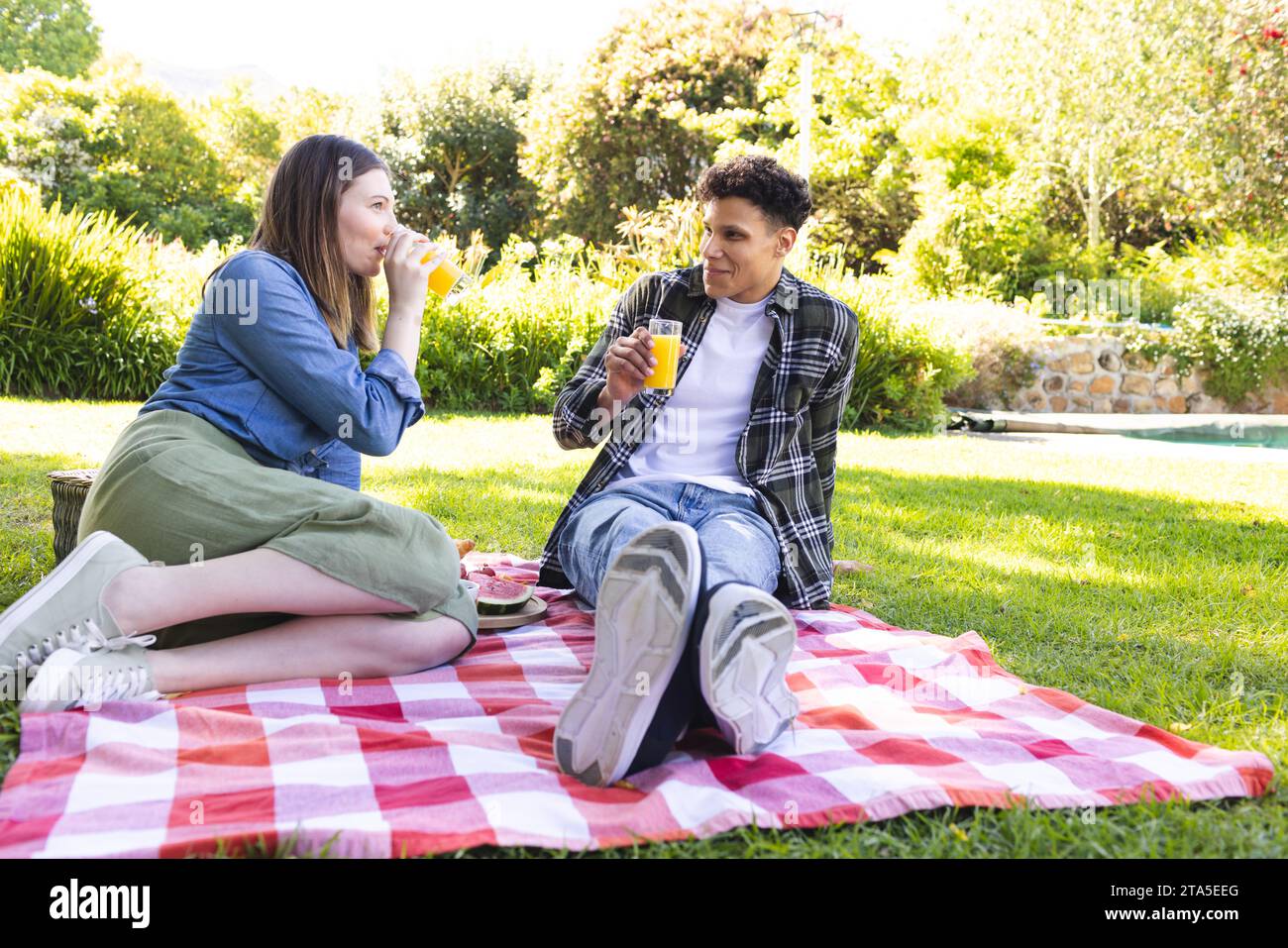 Glückliches, vielfältiges Paar, das auf einer Decke sitzt, Picknick macht, Saft im sonnigen Garten trinkt, Kopierraum. Draußen, Ausdruck, Freizeit, Liebe, Zweisamkeit, Stockfoto