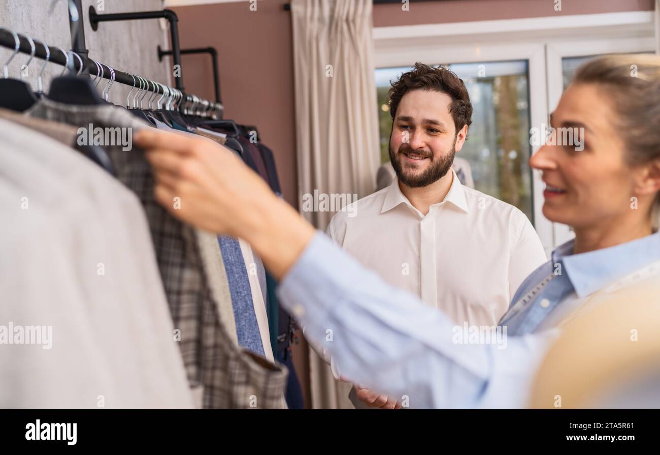 Ein Schneider hält eine karierte Jacke für einen lächelnden männlichen Kunden in einem Hochzeitsgeschäft hoch Stockfoto