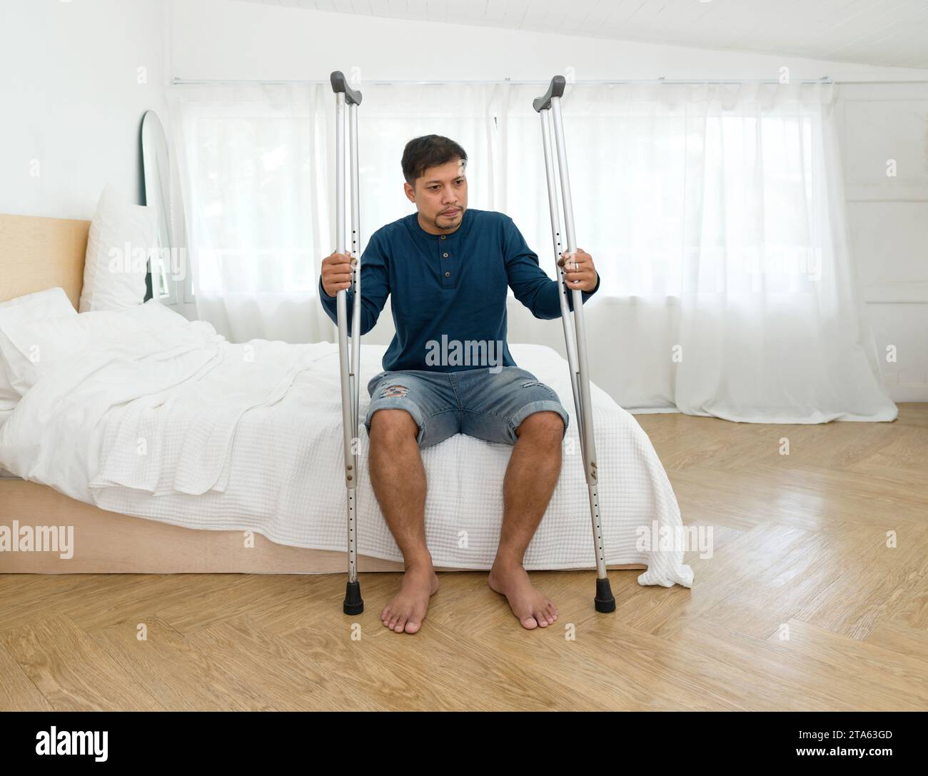 Ein Mann ruht auf einem Bett, sieht besinnlich aus, mit zwei Krücken in der Nähe, was auf eine jüngste körperliche Not hindeutet. Stockfoto