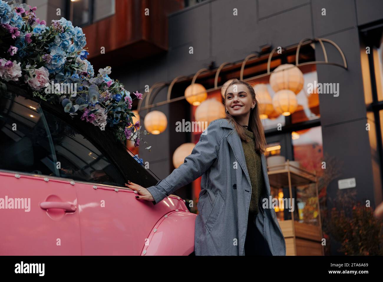 Junge glückliche lächelnde Frau, die neben einem rosa Auto steht, das mit Blumengirlanden auf der Stadtstraße vor dem Hintergrund des Gebäudes dekoriert ist. Nahansicht. Stockfoto