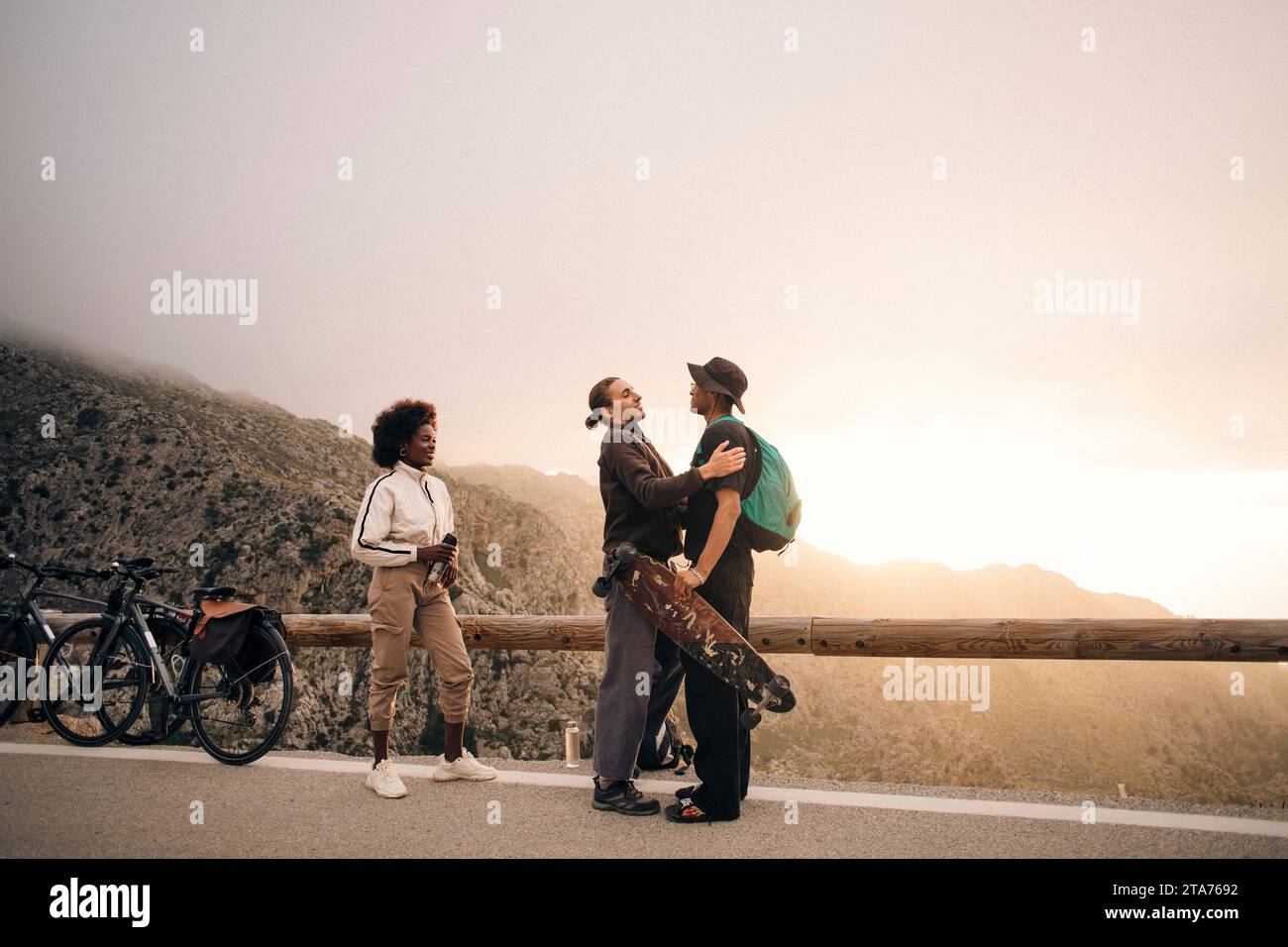 Junge männliche und weibliche Freunde grüßen einander, während sie in der Nähe eines hölzernen Leitplanks auf der Straße gegen den Himmel stehen Stockfoto