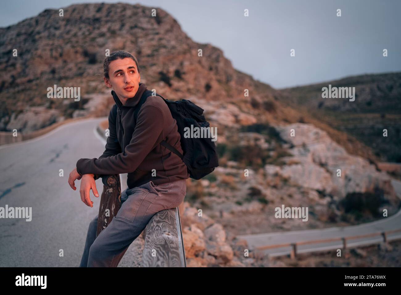 Junger Mann, der Berge erforscht, während er bei Sonnenuntergang auf Geländer sitzt Stockfoto