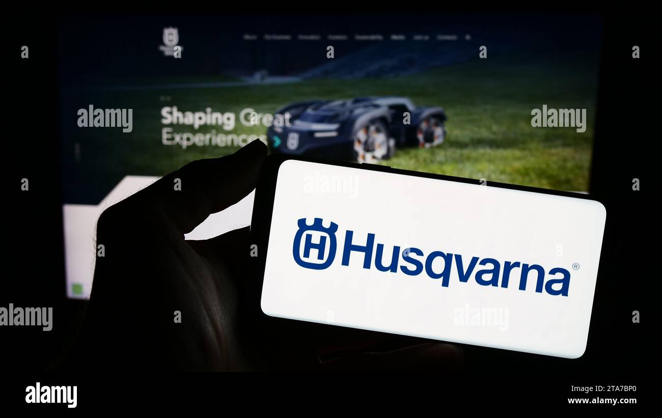 Person mit Smartphone und Logo des schwedischen Stromversorger Husqvarna ab vor der Website. Schwerpunkt auf dem Display des Telefons. Stockfoto