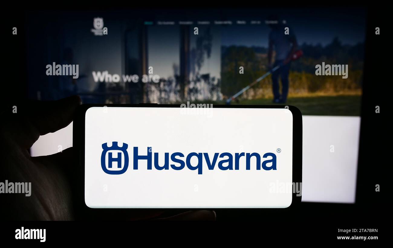 Person, die ein Mobiltelefon mit dem Logo des schwedischen Energieunternehmens Husqvarna ab vor der Webseite hält. Schwerpunkt auf dem Display des Telefons. Stockfoto