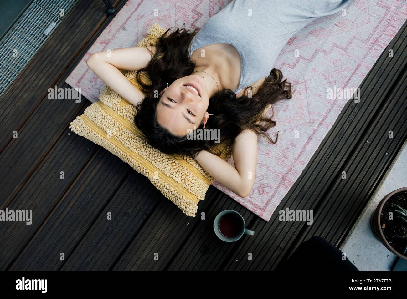 Direkt über dem Blick auf eine lächelnde Frau, die sich mit Händen hinter dem Kopf auf dem Teppich auf dem Balkon liegend Stockfoto