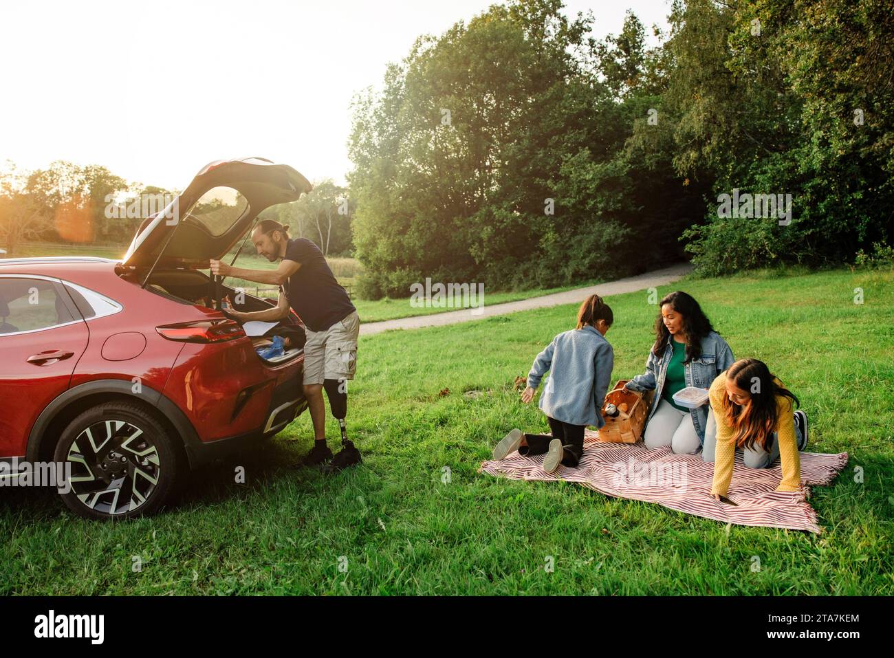 Die Familie bereitet sich auf ein Picknick vor, während sie auf dem Gras in der Nähe des Parkplatzes sitzt Stockfoto