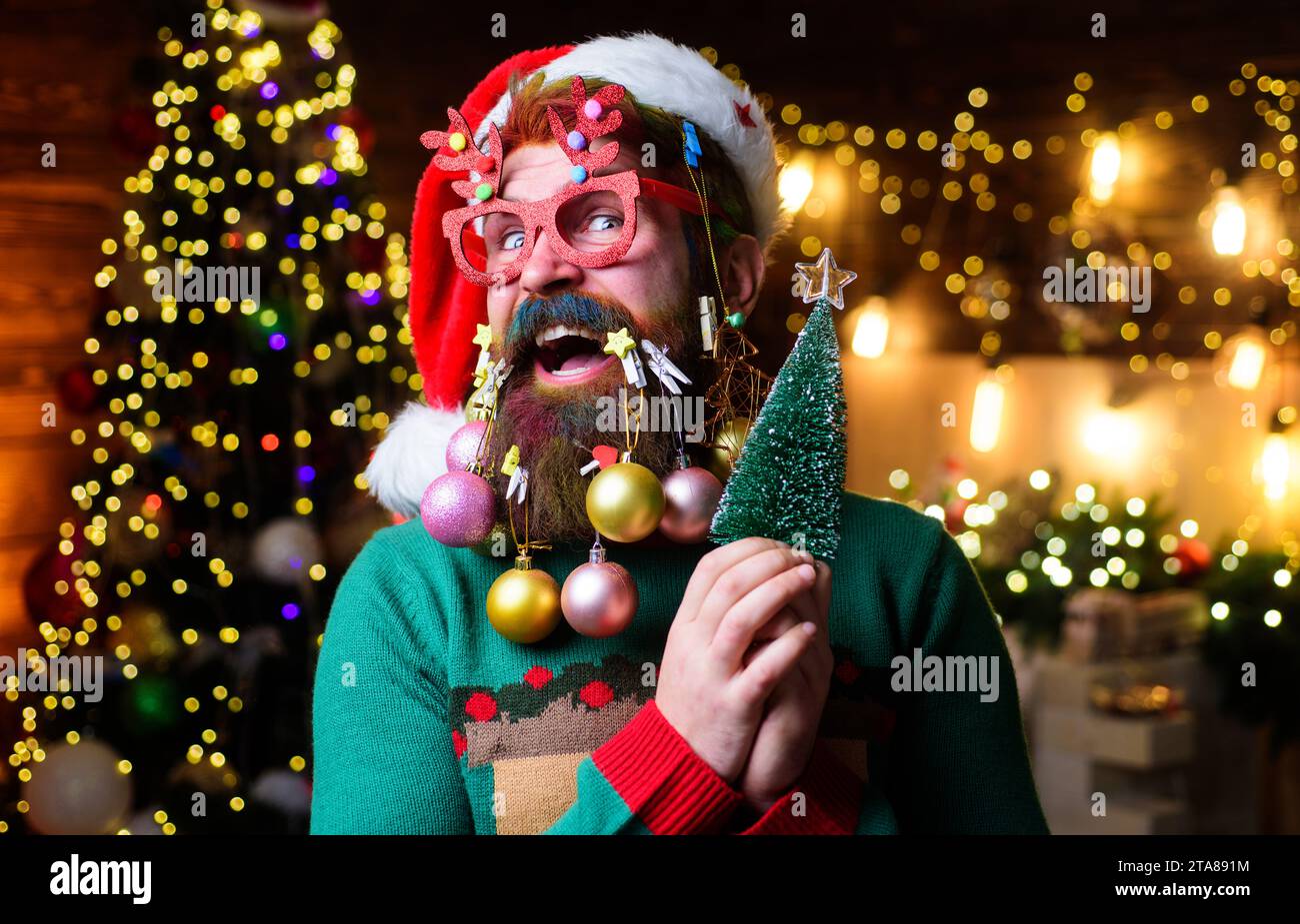 Glücklicher Weihnachtsmann mit Neujahrsbällen im Bart hält einen kleinen Weihnachtsbaum. Weihnachtsbart. Bärtiger Kerl in Partybrille und Weihnachtsmann-Hut mit wenig Stockfoto