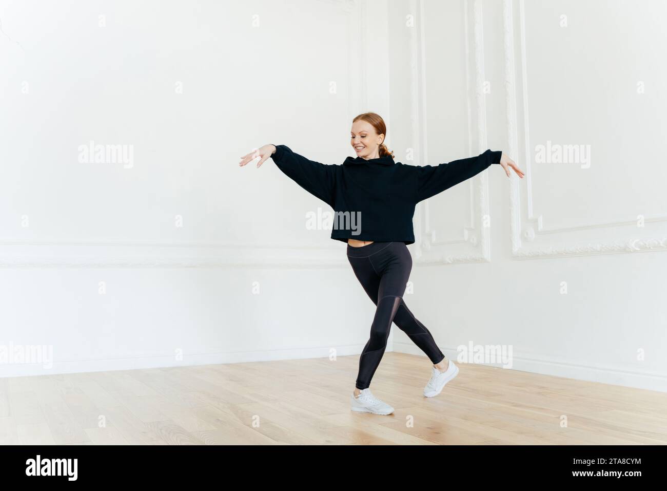 Energiegeladene Frau, die in einem schwarzen Crop-Top und Leggings tanzt, geräumigen weißen Raum, der Freude und Bewegungsfreiheit ausdrückt Stockfoto