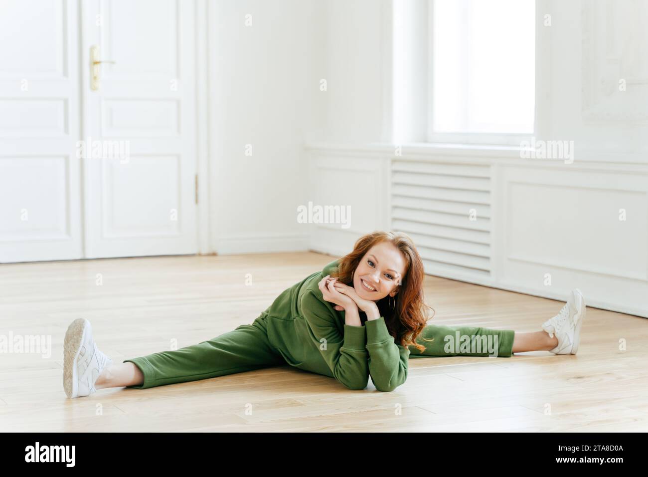 Entspannte Frau in grüner Loungewear, die auf Holzboden liegt, elegante weiße Innenausstattung, friedlicher Ausdruck und bequeme Pose Stockfoto