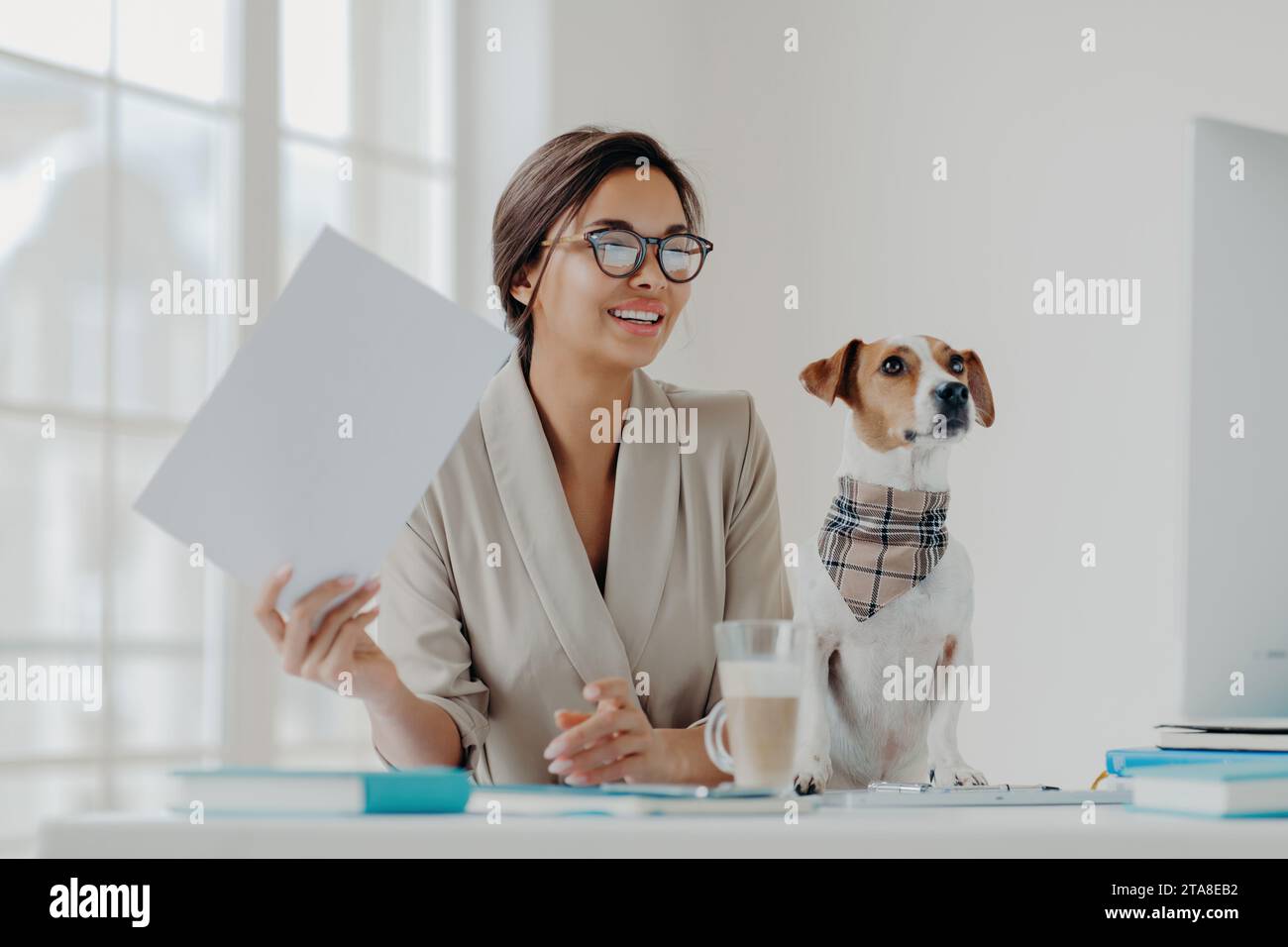 Lächelnde Profi-Frau, die Dokumente mit ihrem aufmerksamen Hund in einem hellen Heimbüro hält und Effizienz verkörpert. Stockfoto