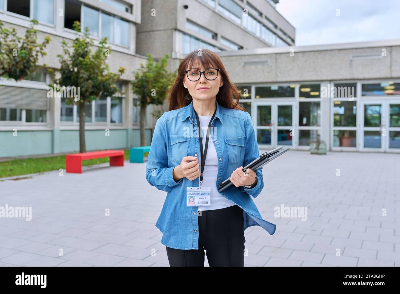 Schullehrerin mittleren Alters mit digitalem Tablet posiert in der Nähe des Schulgebäudes Stockfoto