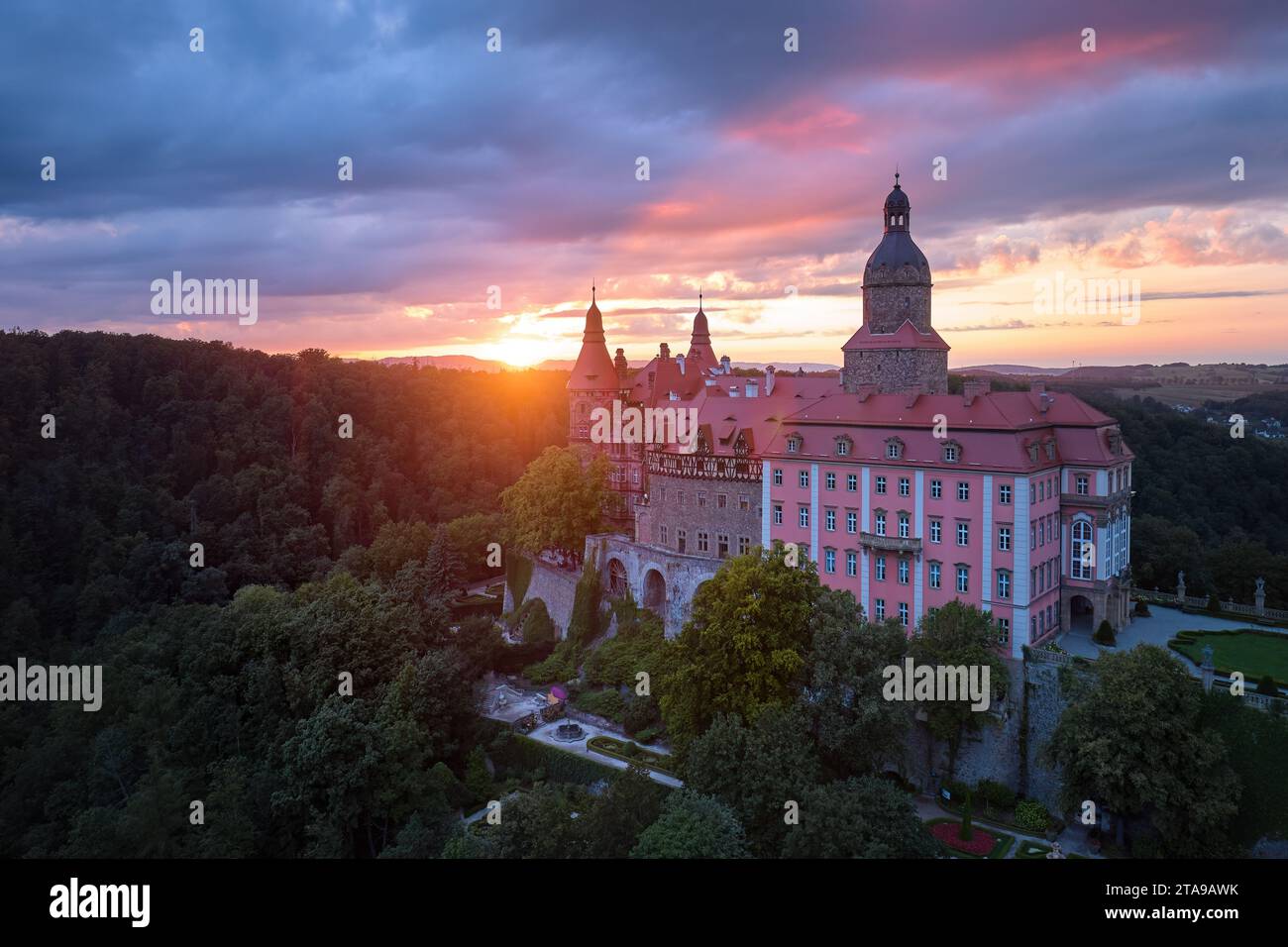 Ein abendlicher Blick aus der Luft auf das rote, sonnendurchflutete Schloss Ksiaz, Schloss Fürstenstein, ein wunderschönes Schloss auf einem Felsen, umgeben von Wald, gegen einen Dram Stockfoto