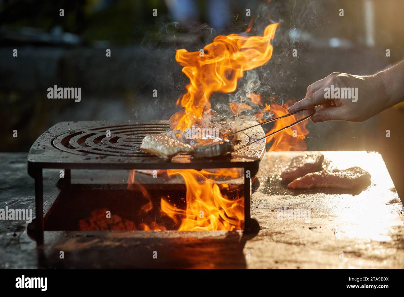 Ein Koch bereitet ein Steak auf einem Grill im Freien zu. Nahaufnahme des Kochens beim Gartenfest, Flammen, geringe Tiefe des Feldes, sehr bunter, verschwommener Hintergrund, Stockfoto