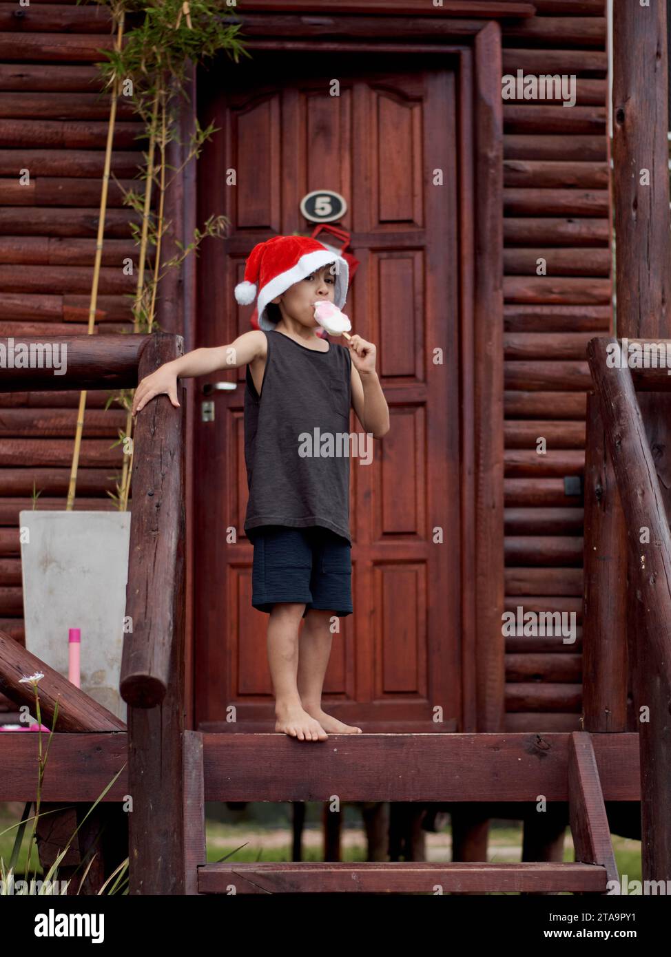 Latein kleiner Junge vor einer Holzhütte, der ein Eis in der Hand hält und Weihnachten im Sommer feiert. Trägt einen roten Weihnachtsmann-Hut und ärmellos Stockfoto