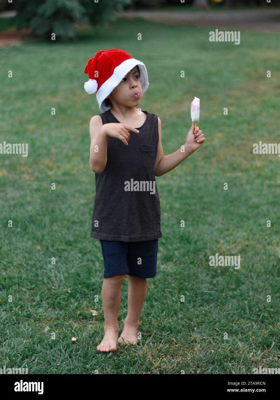 Entspannter Latin Boy, der im Sommer ein Eis genießt und Weihnachten vorwegnimmt, trägt Shorts, ein Tanktop und einen Weihnachtsmann-Hut auf dem Rasen seines Gartens. Stockfoto