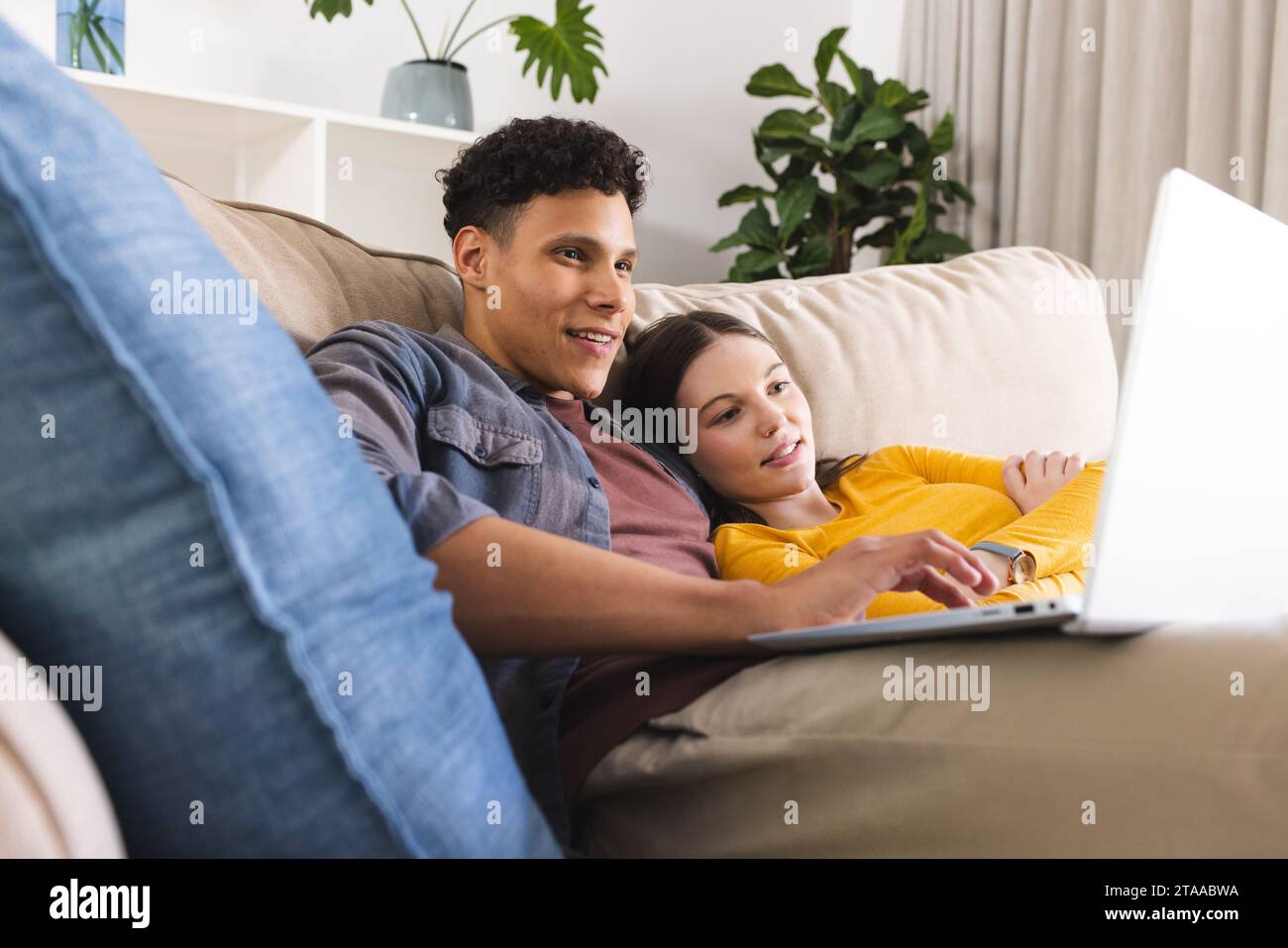 Glückliches, vielfältiges Paar, das auf der Couch liegt und im Wohnzimmer zu Hause einen Laptop benutzt, Kopierraum. Liebe, Zweisamkeit, Technologie, Kommunikation und häusliches Leben Stockfoto