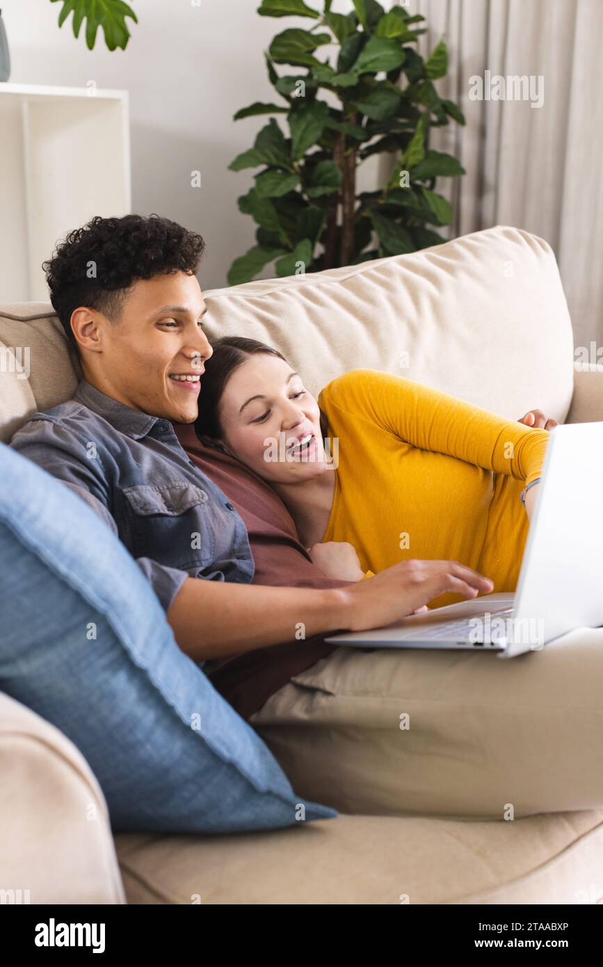 Glückliches, vielfältiges Paar, das auf der Couch liegt und im Wohnzimmer zu Hause einen Laptop benutzt, Kopierraum. Liebe, Zweisamkeit, Technologie, Kommunikation und häusliches Leben Stockfoto