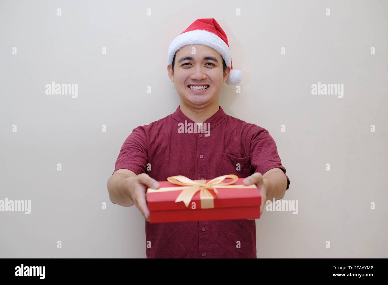 Der junge asiatische Weihnachtsmann schenkt der Kamera eine rote Geschenkbox mit einem Lächeln. Isolierter beigefarbener Hintergrund. Weihnachtskonzept. Stockfoto