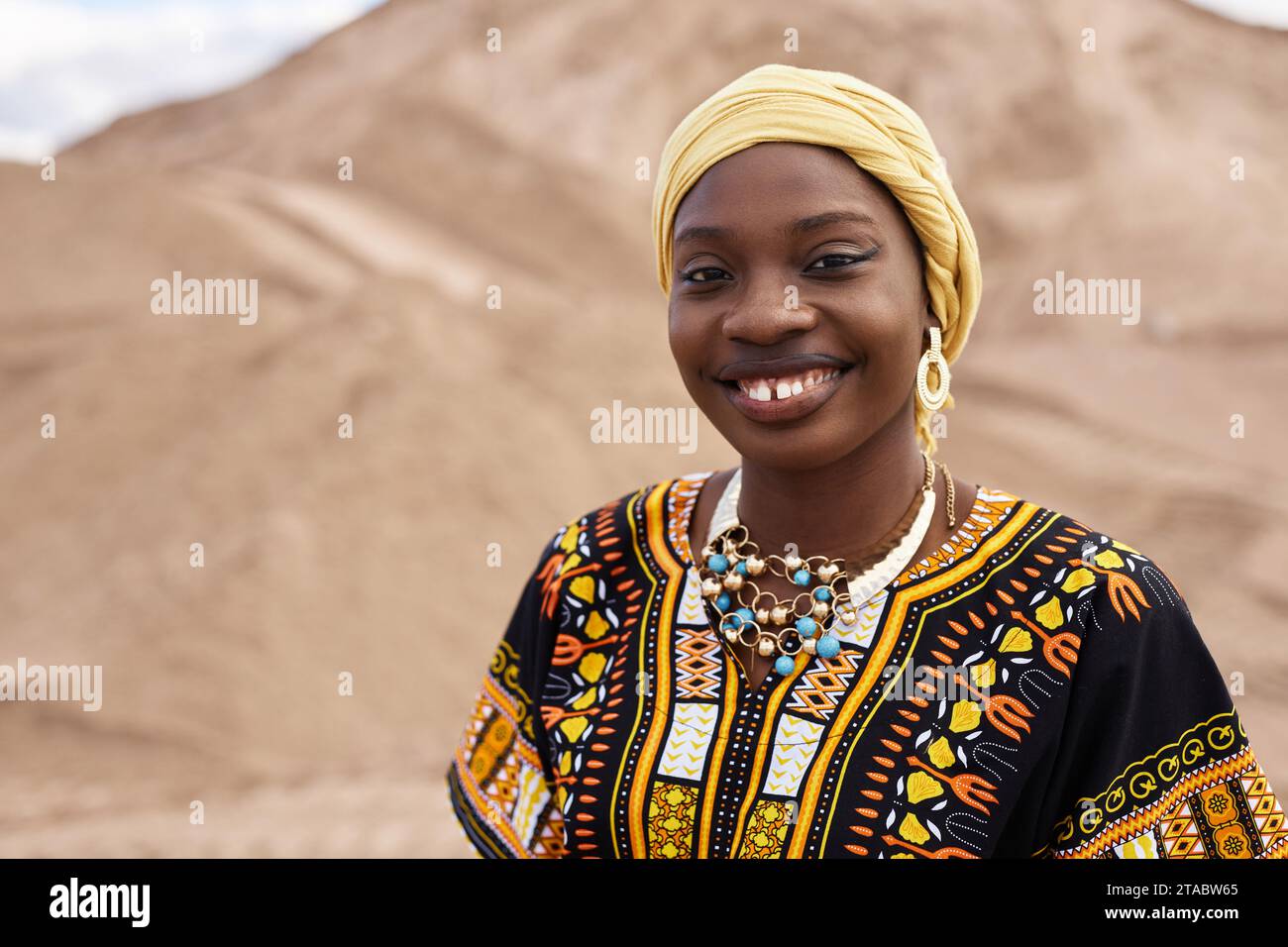 Porträt einer jungen Schwarzen Frau, die ein Outfit mit traditionellen afrikanischen Ornamenten trägt, während sie in der Wüste posiert und lächelnd im Kopierraum erscheint Stockfoto