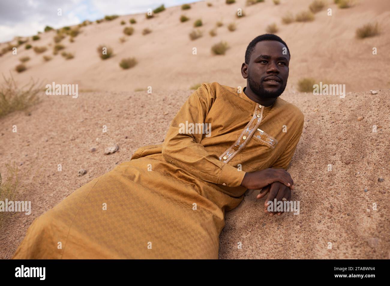 Porträt eines afroamerikanischen Mannes in langem Kaftan-Kleid, der auf Sanddünen in der Wüste liegt und wehmütig wegblickt Stockfoto