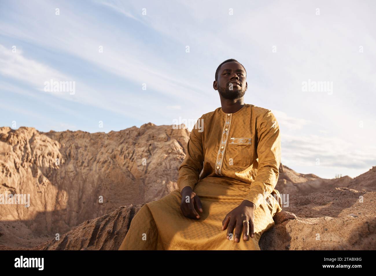Dramatisches Porträt des jungen Schwarzen Mannes, der lange Kaftan trägt und Sanddünen sitzt und in die Wüste blickt, Kopierraum Stockfoto
