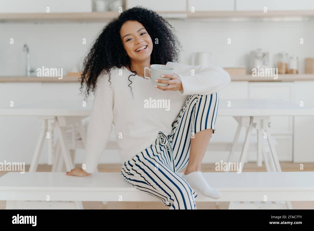 Lächelnde schwarze Frau in einem weißen Pullover, hält einen Becher, sitzt an einem Küchentisch Stockfoto