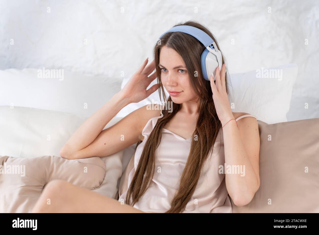 Kaukasische junge Frau mit Kopfhörern hört Musik, während sie im beigefarbenen Seidenschlafanzug im Bett sitzt Stockfoto