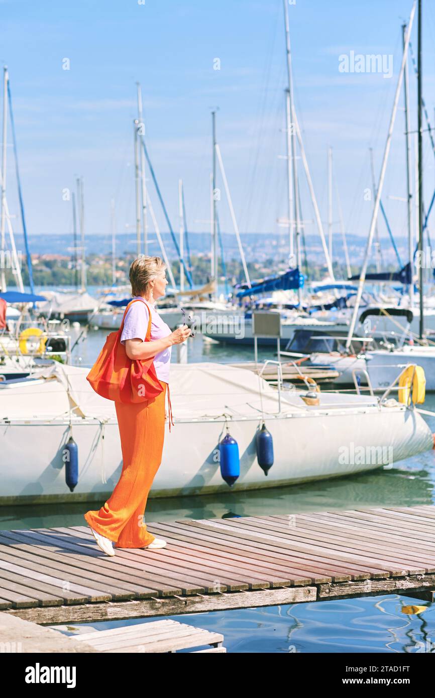 Outdoormodeporträt von mittlerem Alter 50 - 55 Jahre alte Frau in bunten Outfit, posiert am See oder Hafen, Reise- und Urlaubsleben Stockfoto