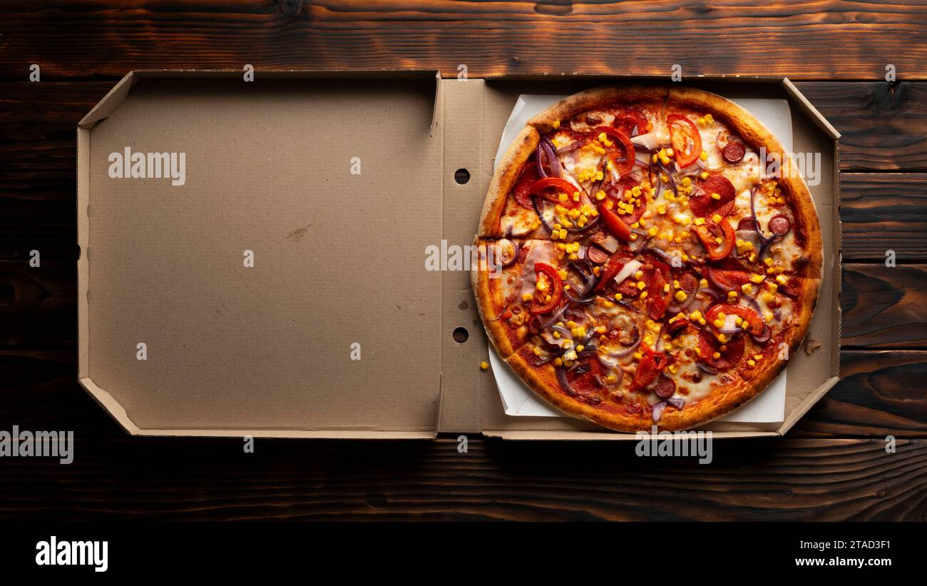 Paprika-Pizza mit Paprika und Zuckermais in offener Kartonbox auf dunklem Holztisch flach liegend mit Kopierraum Stockfoto
