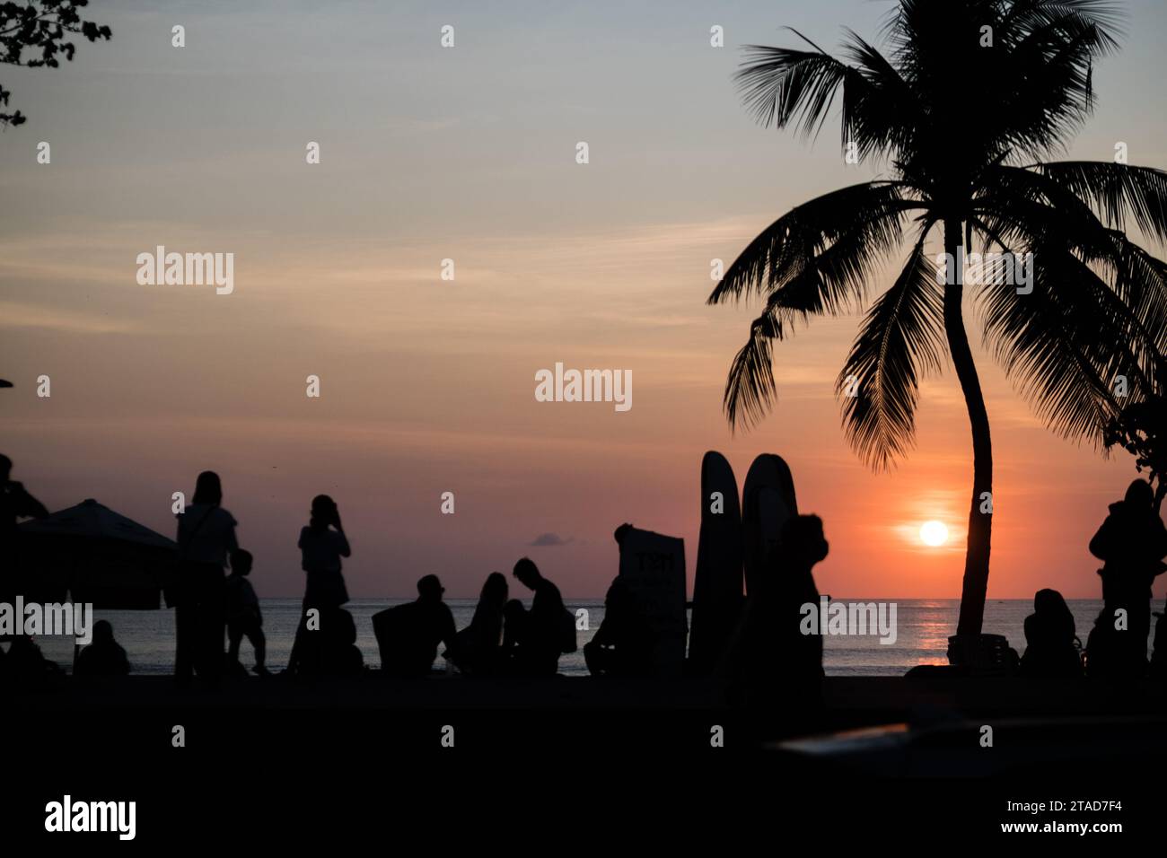 Silhouetten von Menschen und eine Palme vor einem pulsierenden Sonnenuntergang an einem tropischen Strand schaffen eine ruhige und malerische Abendszene am Kuta Beach, Bali Stockfoto