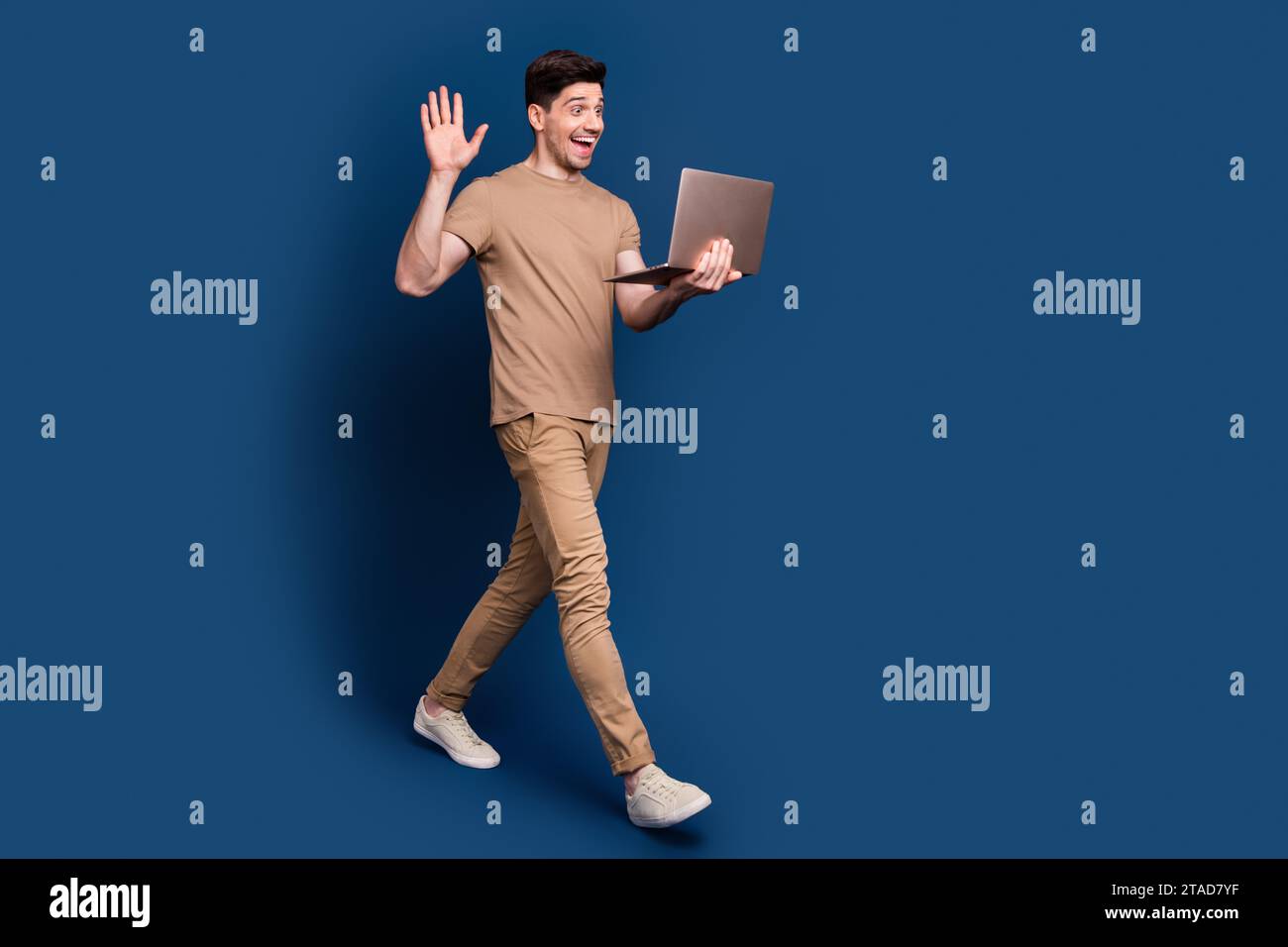 Foto in voller Länge des fröhlichen Mannes tragen beige stilvolles Outfit winkende Handfläche Sagen Sie hallo am Laptop auf Webcam isoliert auf dunkelblauem Hintergrund Stockfoto