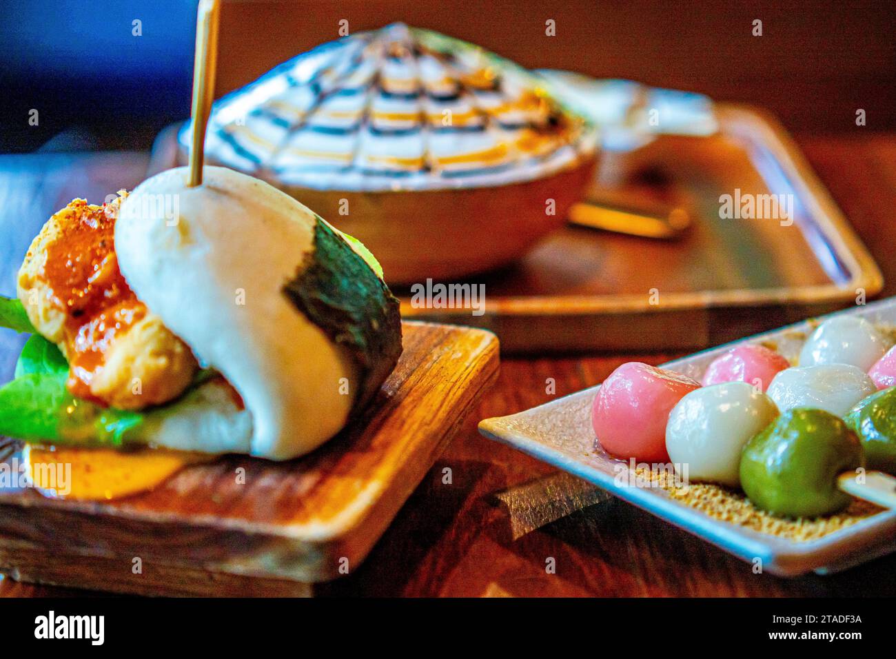 Trendige Café-Tischplatte mit knusprigem Huhn-bao-Brötchen, dreifarbigen Dancobießen und einem rasierten Eisdessert mit Schokoladennebel. Stockfoto