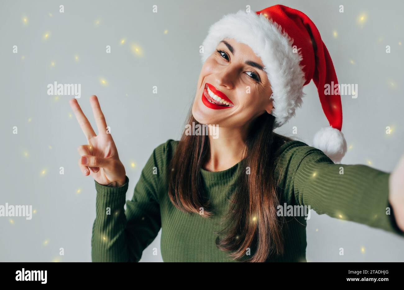 Nahaufnahme Porträt einer hübschen süßen jungen Frau mit Friedensgeste, Weihnachtsmann Hut und Selfie isoliert über grauem Hintergrund. Glücklich Stockfoto