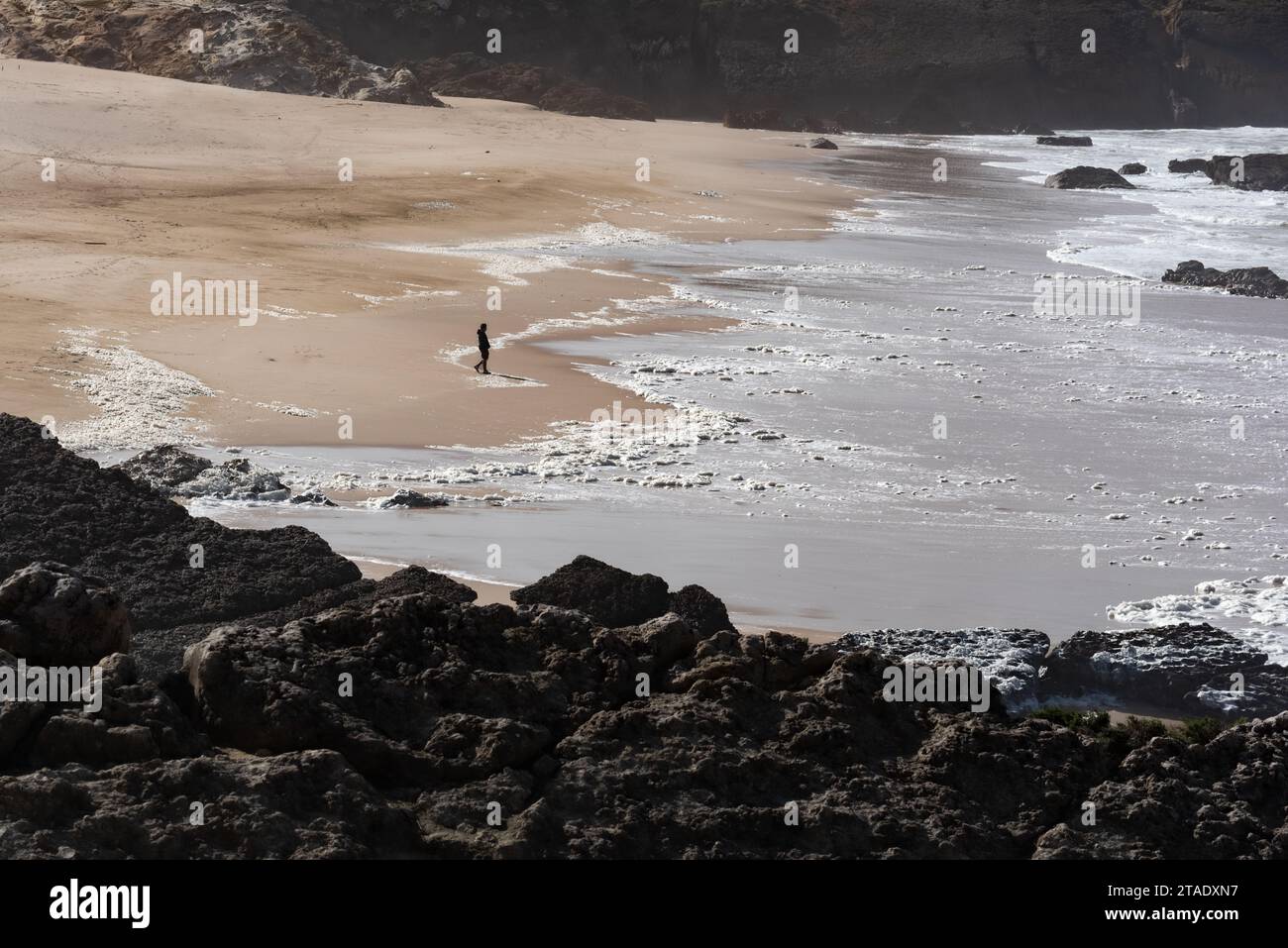 Die Silhouette einer einzelnen Person, die sich in der Nebensaison in Cascais in Portugal an einem leeren Sandstrand von Cresmina dem Meeresrand nähert Stockfoto